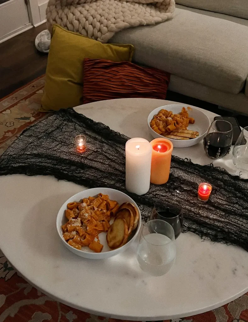Two bowls of pasta and glasses of red wine on a round marble coffee table with lit candles, a chunky knit blanket and mustard and rust cushions