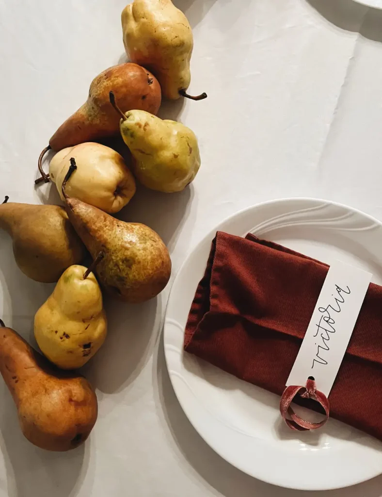White table setting with a rust napkin, handwritten place card and pears arranged along the tablecloth as a centerpiece