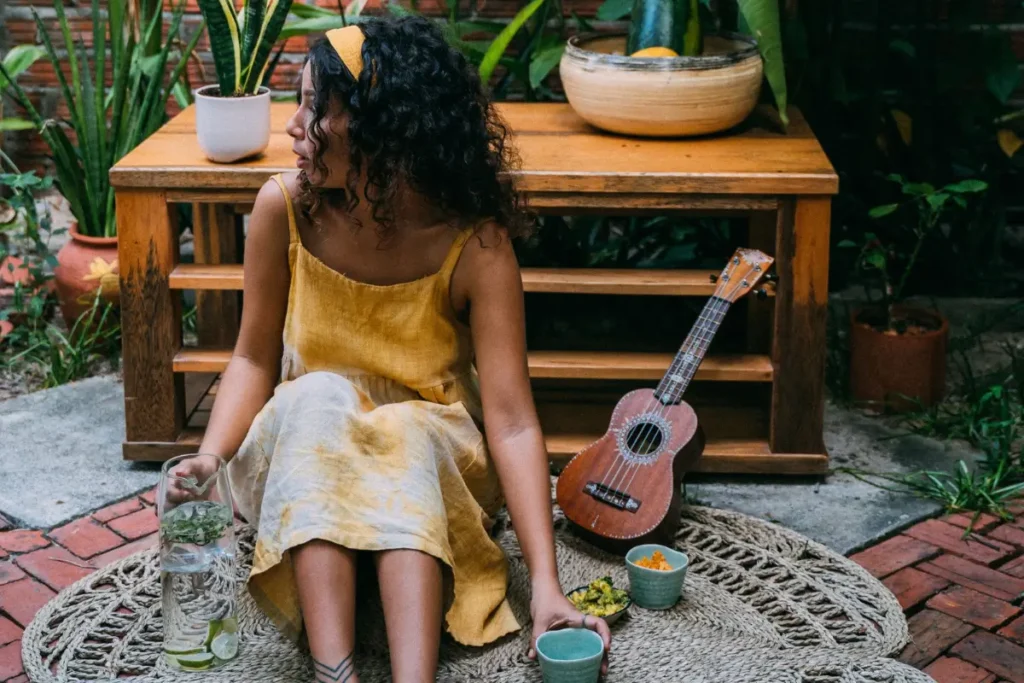 Woman in a yellow dress sitting with a ukulele in a lush indoor setting, enjoying a peaceful moment.