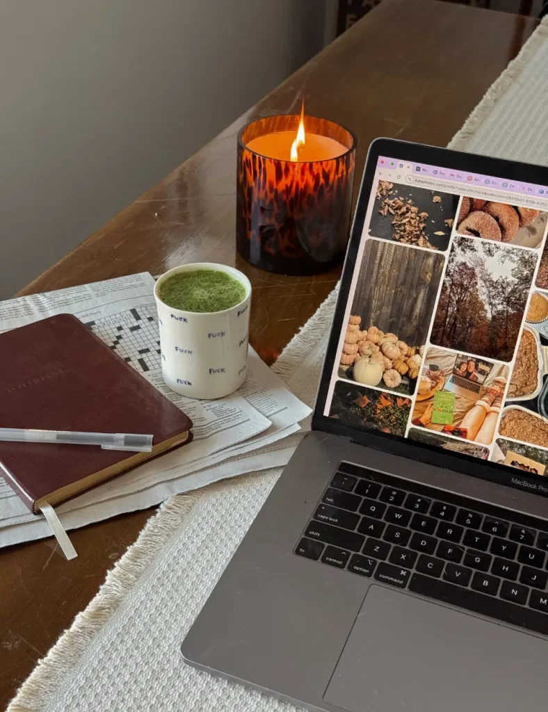 Wooden table with a leather journal and pen, a matcha latte, a lit amber candle, a newspaper crossword and an open laptop