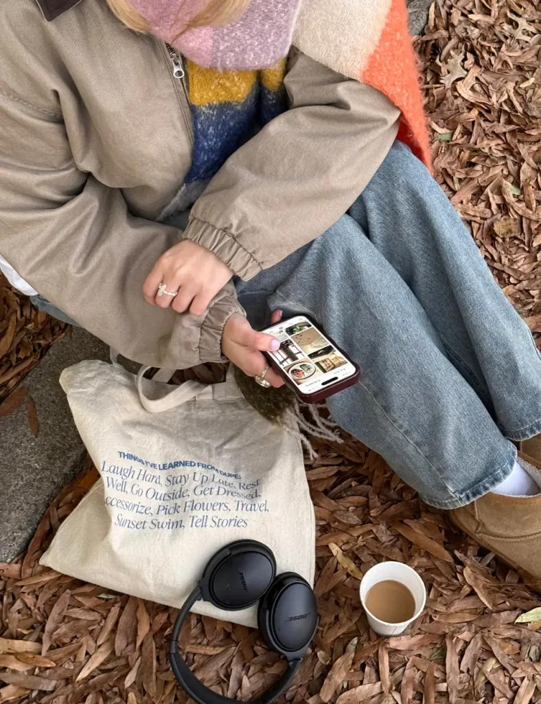 Person in a beige jacket and mohair scarf sitting on a blanket in fallen leaves with a phone, tote bag, headphones and a coffee cup