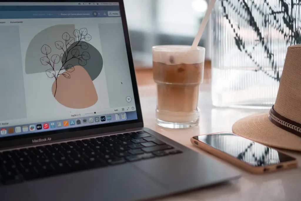 Botanical wall art design open in Canva on a laptop beside an iced coffee and a straw hat.