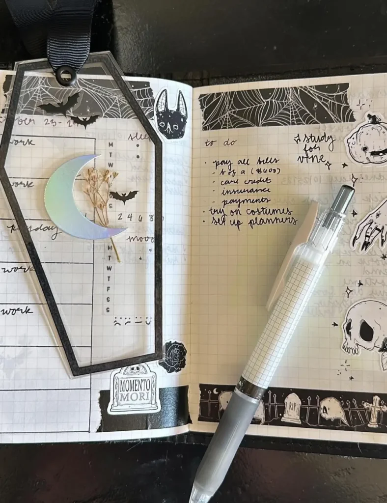 Coffin-shaped cutout bookmark tracker with a moon charm and Memento Mori tombstone sticker in a Halloween journal