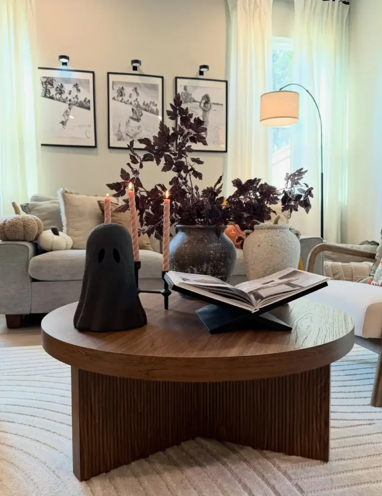 Round wood coffee table styled with a black ghost candle holder, pink taper candles, dark foliage in a stone vase and an open book on a stand