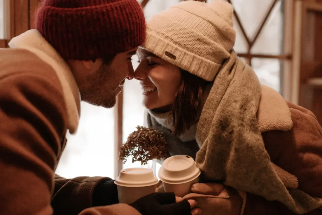 A cozy couple enjoys coffee indoors, touching noses and smiling in winter attire.