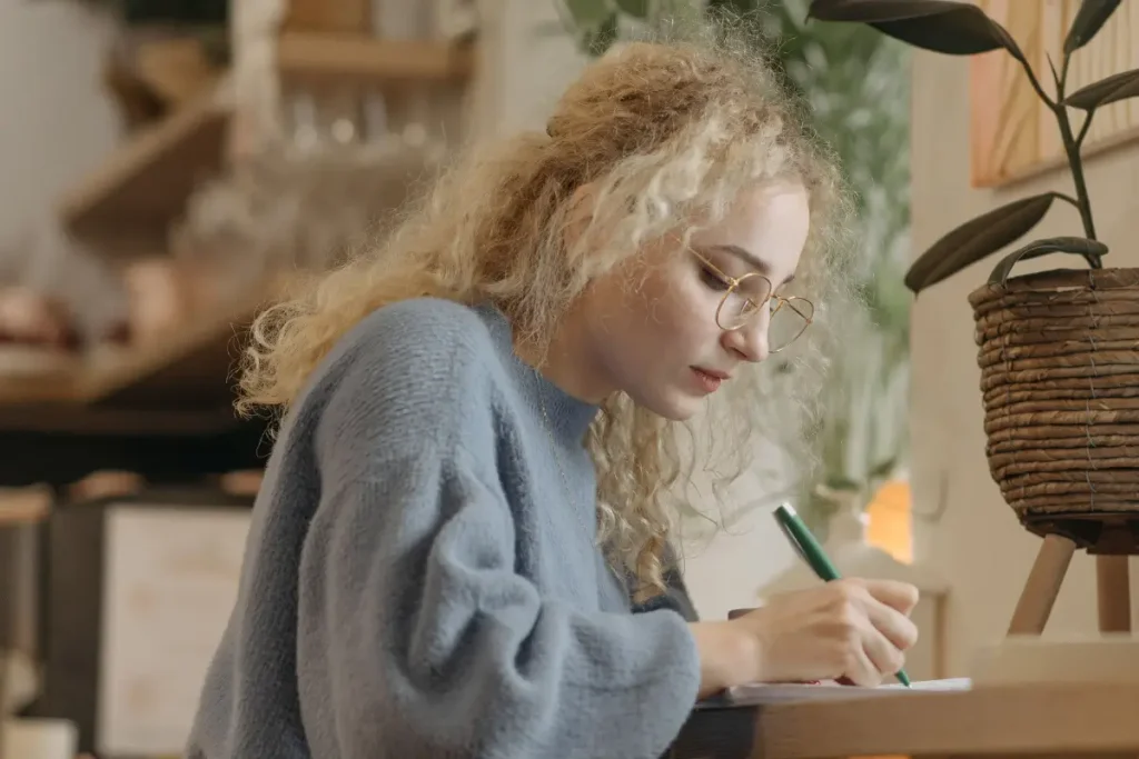 A young woman wearing a blue sweater writes at a table in a cozy, plant-filled indoor setting.