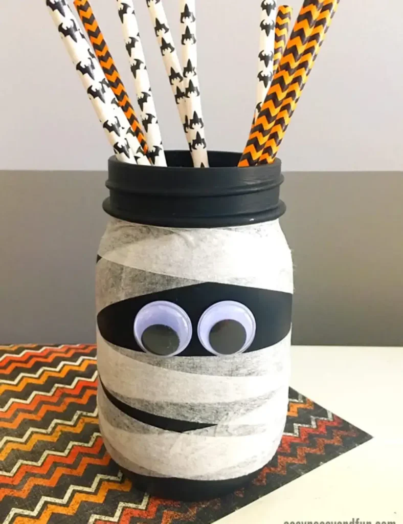 Black mason jar wrapped in white tape strips to look like a mummy, with googly eyes glued near the top