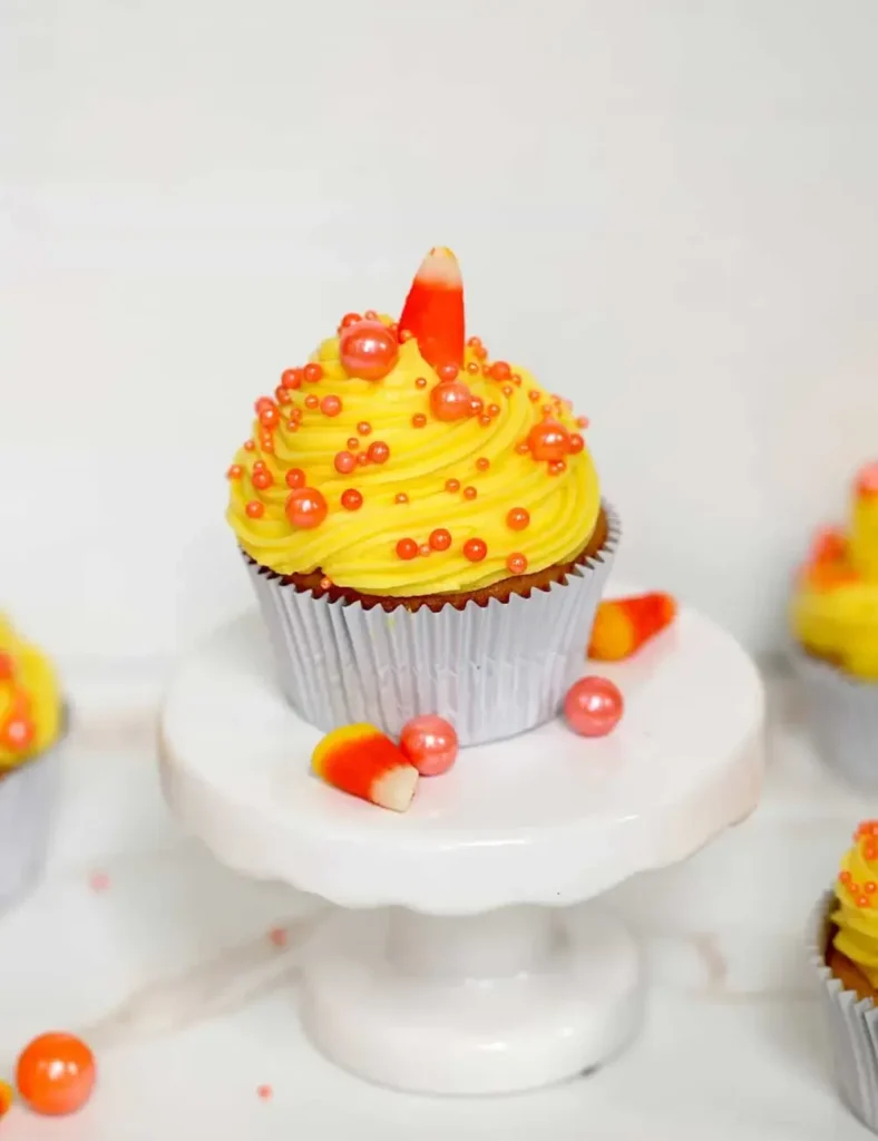 Candy corn cupcake with yellow frosting, orange sprinkles and a candy corn topping on a white cake stand
