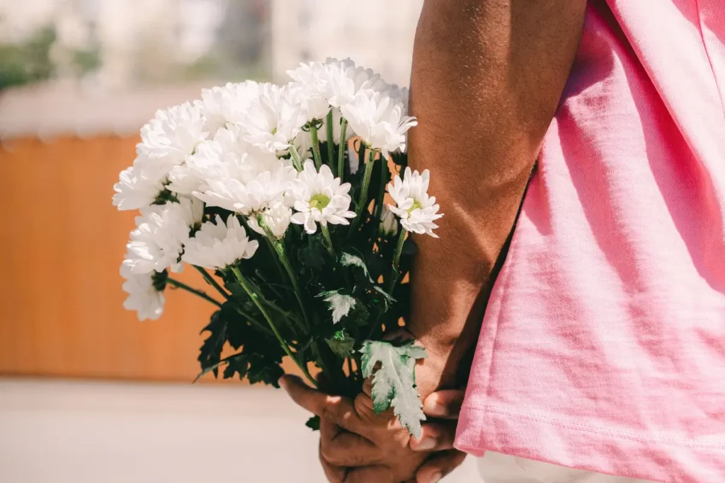 Close-up of a man in pink shirt holding a bouquet of white flowers behind his back, outdoors