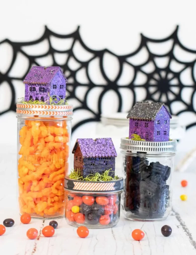 Mason jars topped with glittery purple and black miniature haunted houses and moss, filled with Halloween candy