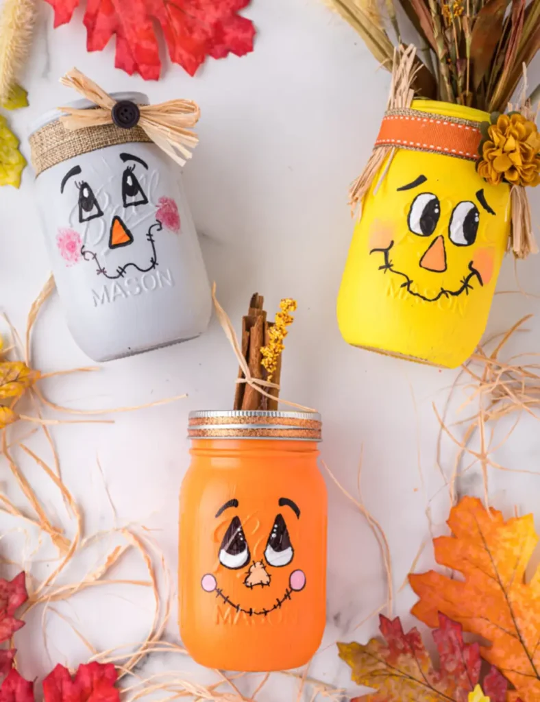 Scarecrow mason jars painted yellow, gray and orange with hand-drawn faces and fall ribbon accents