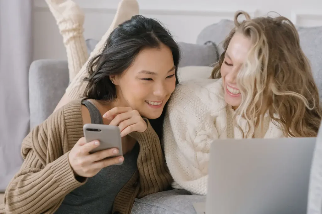 Two women relaxing on the floor while engaging with digital devices, indoors and smiling.