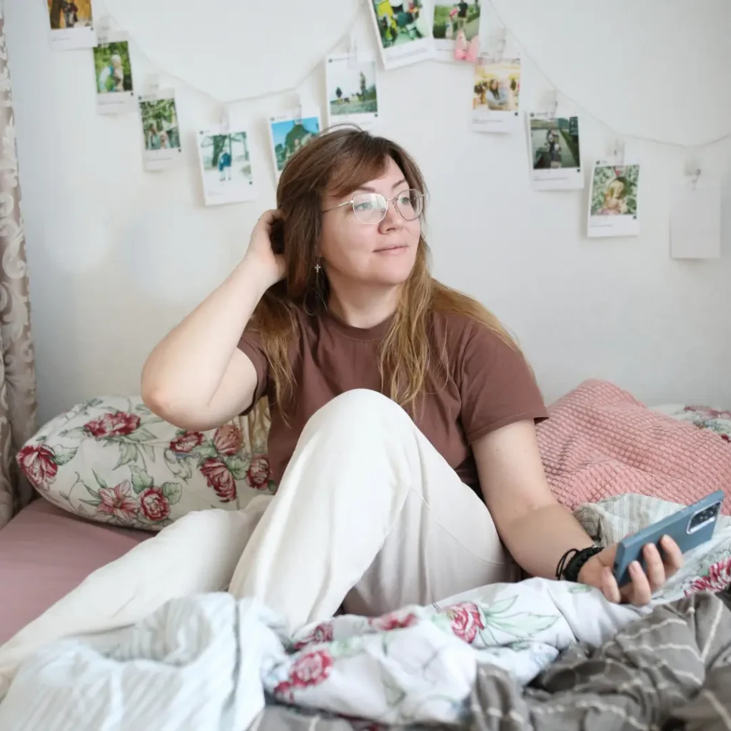 A woman comfortably sitting in bed holding a smartphone, surrounded by cozy decor