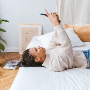 African American woman lying on bed using smartphone in cozy, stylish bedroom.