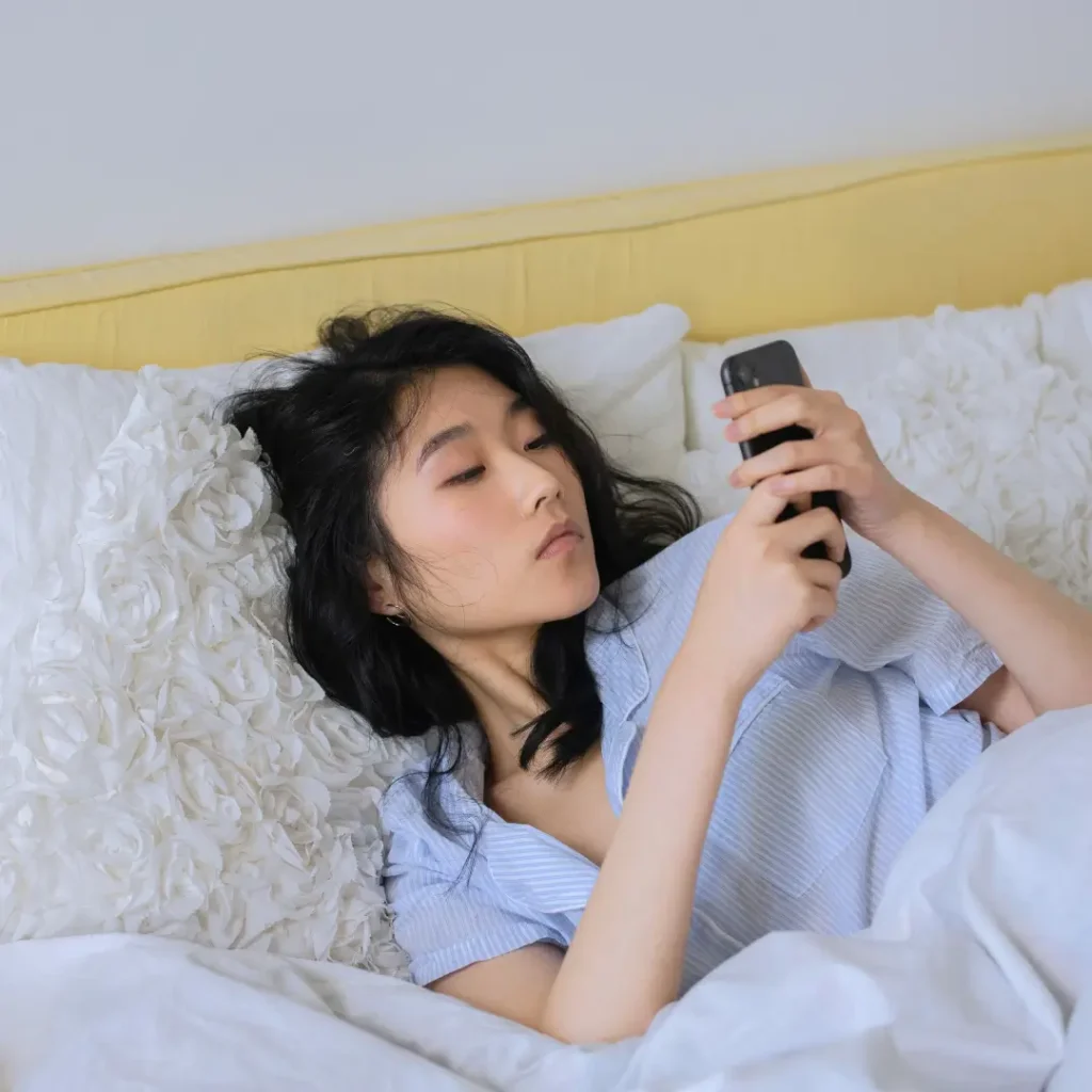 Asian woman comfortably lying in bed, using her phone in a serene bedroom setting