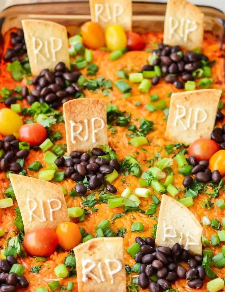 Baked Halloween taco dip decorated with tortilla tombstones reading _RIP_ among black beans and tomatoes