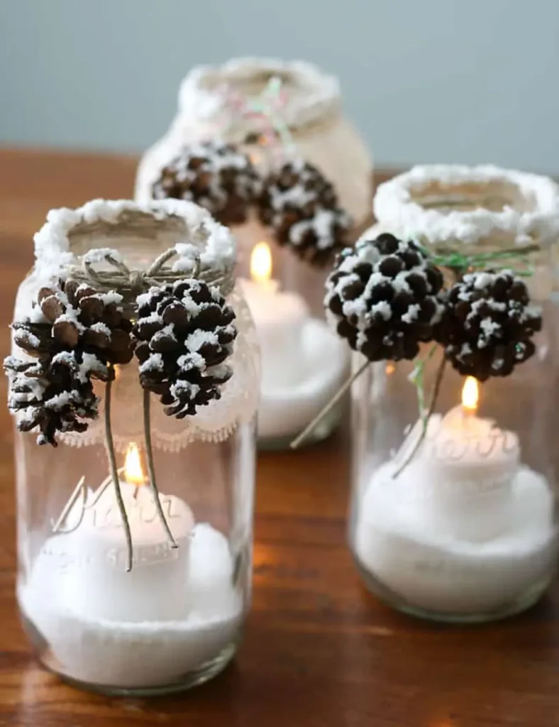Glass jar luminaries with lace and twine around the rim, snow-dusted pinecones on top and a glowing candle inside.