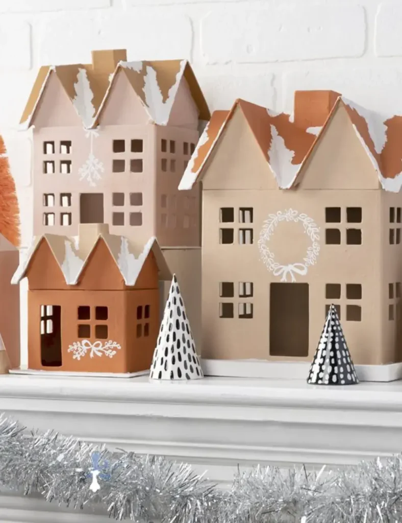 Neutral paper mache Christmas village with stenciled houses, snow paste roofs, shadow box trees and painted cone trees on a mantel