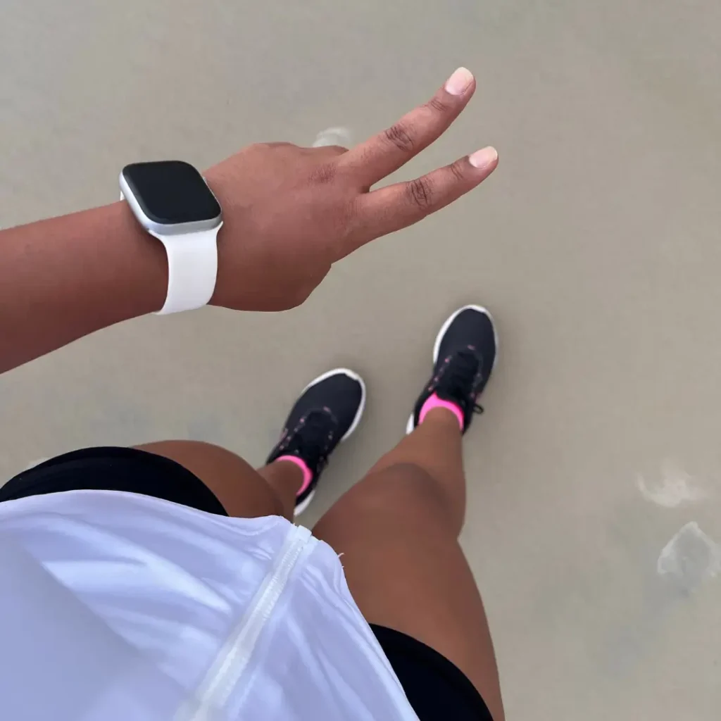 Overhead view of a woman’s legs on wet sand in black and pink sneakers, with her hand making a peace sign and wearing a white smartwatch.