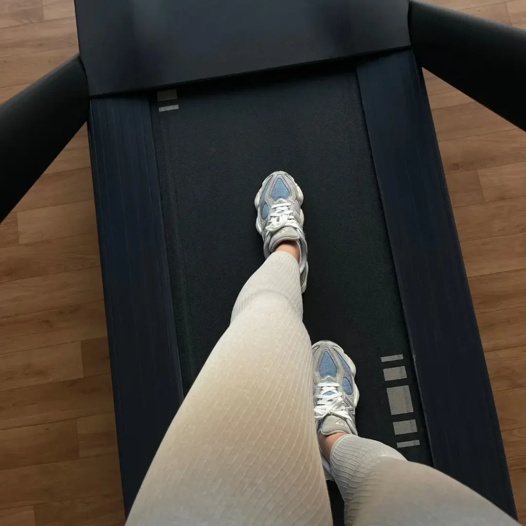 Top-down view of feet in chunky gray and blue sneakers and cream ribbed leggings walking on a treadmill over a wood floor.