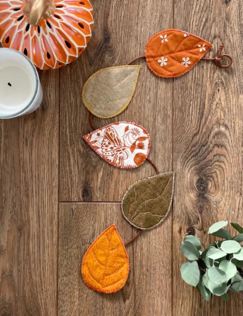 Vertical garland of five stitched fabric autumn leaves in warm colors hanging from a ribbon on a fireplace mantel