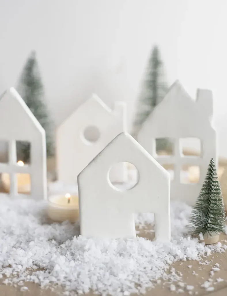 White air dry clay Christmas village houses with cut-out windows and doors, standing on a mantel beside fairy lights