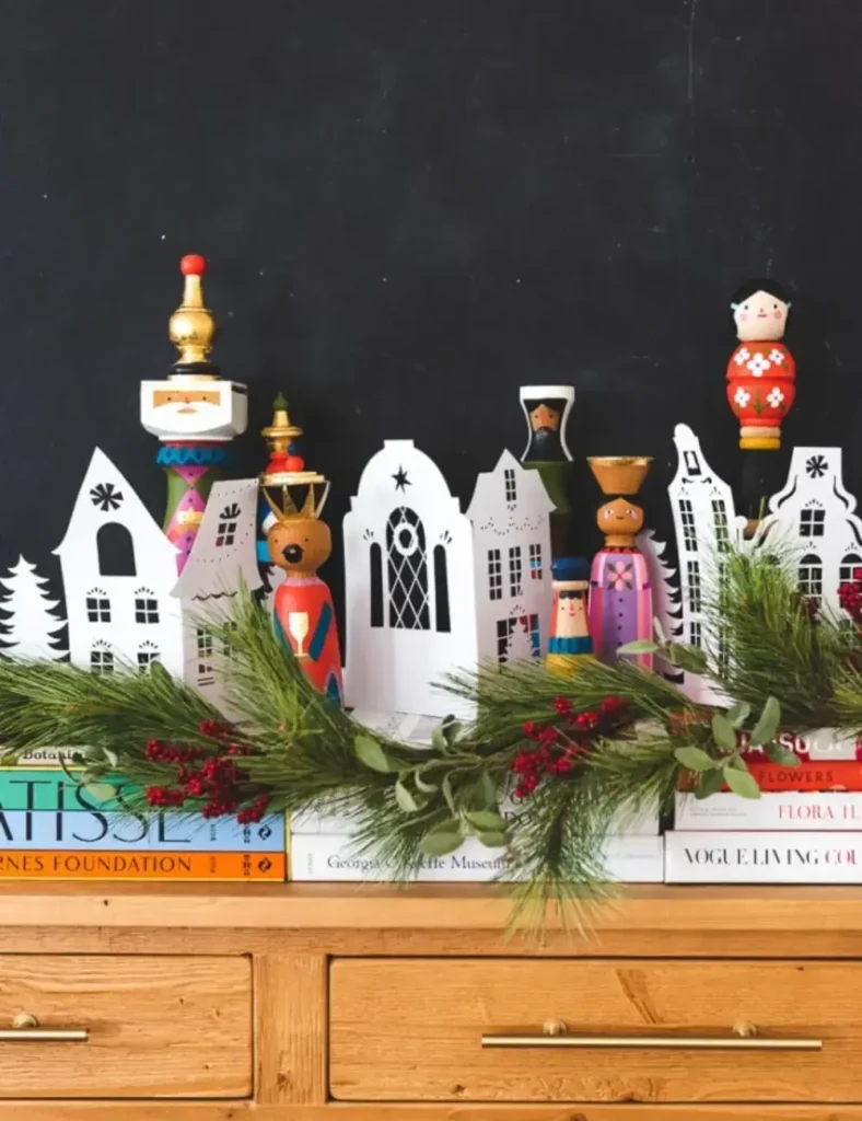 White paper accordion Christmas village standing on a mantel with bottle brush trees, cotton ball clouds and twinkle lights