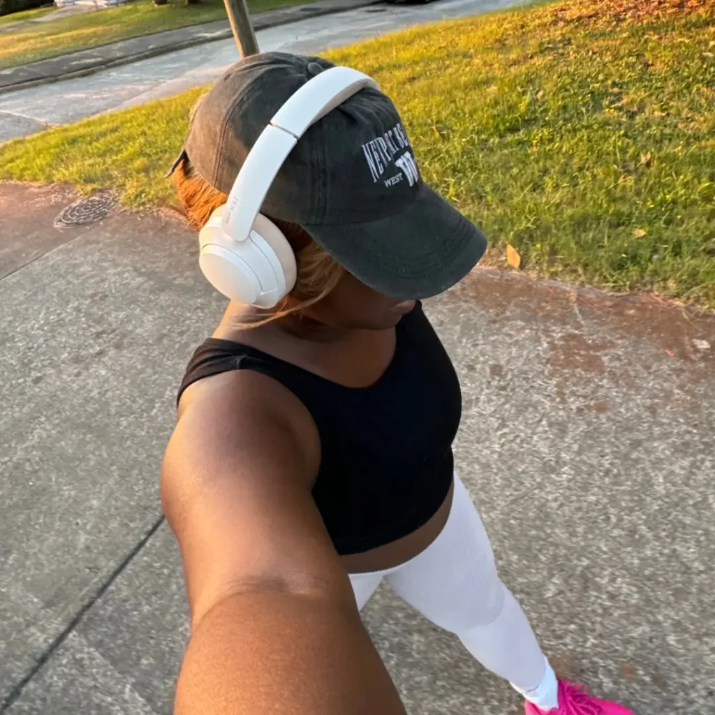 Woman on a sunny sidewalk walk wearing a black cap, white over-ear headphones, black crop top, white leggings, and pink sneakers.